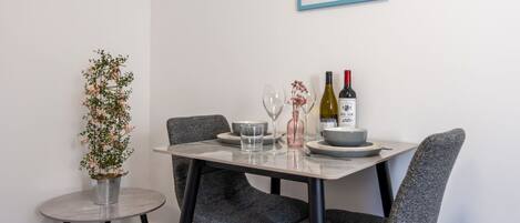 A dining area with a table set for two, featuring a framed picture of a lighthouse, a potted plant, and a bottle of wine.