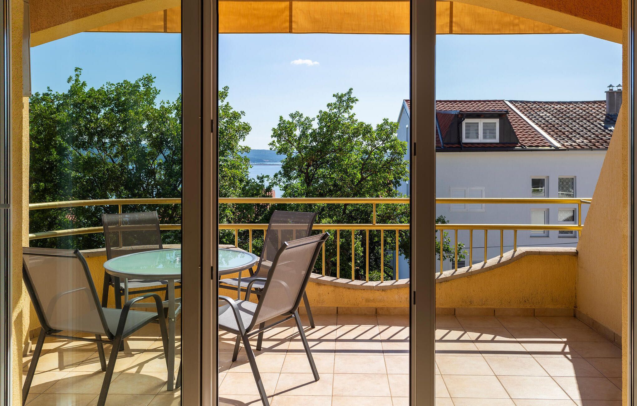 Een balkon met een glazen tafel en stoelen, met uitzicht op een woonwijk met bomen en een gebouw met pannendak.