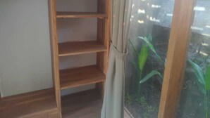 A corner of a room with a wooden shelf unit against a wall, a window with a curtain, and a view of a brick wall and greenery outside. - Keramas House (Gianyar)