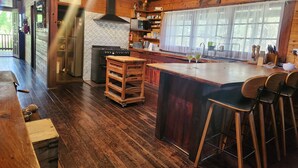 A rustic kitchen with wooden beams, a large island with bar stools, a stove, and a window with sheer curtains. - Secluded rustic cottage charm, opposite river walk, close to town. Pet friendly! (Bridgetown)