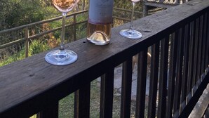A bottle of wine and two glasses on a wooden deck with a view of trees and a wooden fence. - Secluded rustic cottage charm, opposite river walk, close to town. Pet friendly! (Bridgetown)