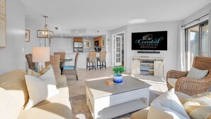 A modern living room with a dining area, kitchen, and a large TV displaying 'Coastal Vacation Rentals'. - Beachfront condo with indoor and outdoor pools! (Hilton Head Island)