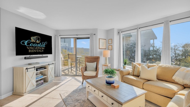 A spacious living room with a large TV, a sofa, a coffee table, and a balcony with a chair. - Beachfront condo with indoor and outdoor pools! (Hilton Head Island)