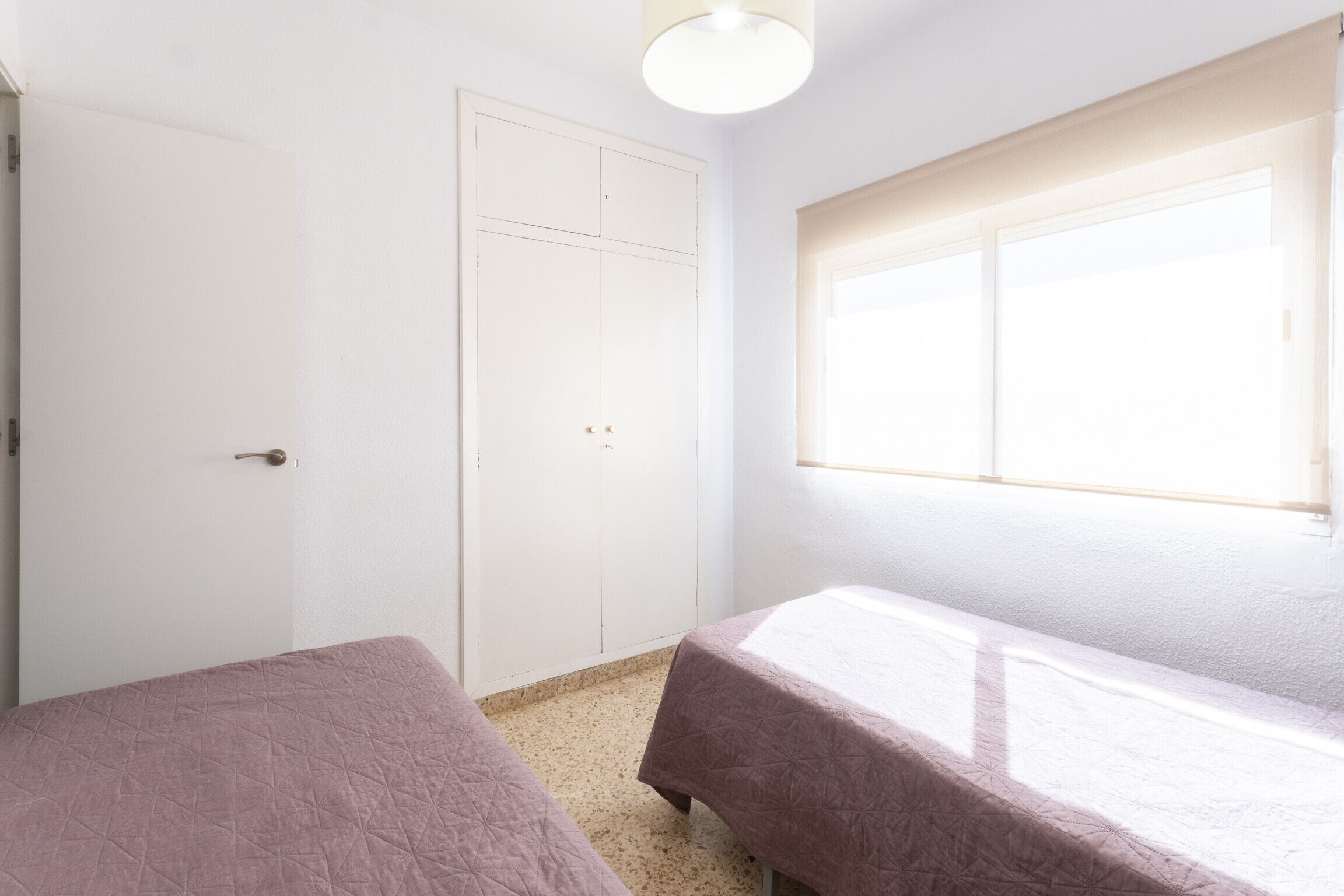 A room with two beds, a white door, and a window with blinds.