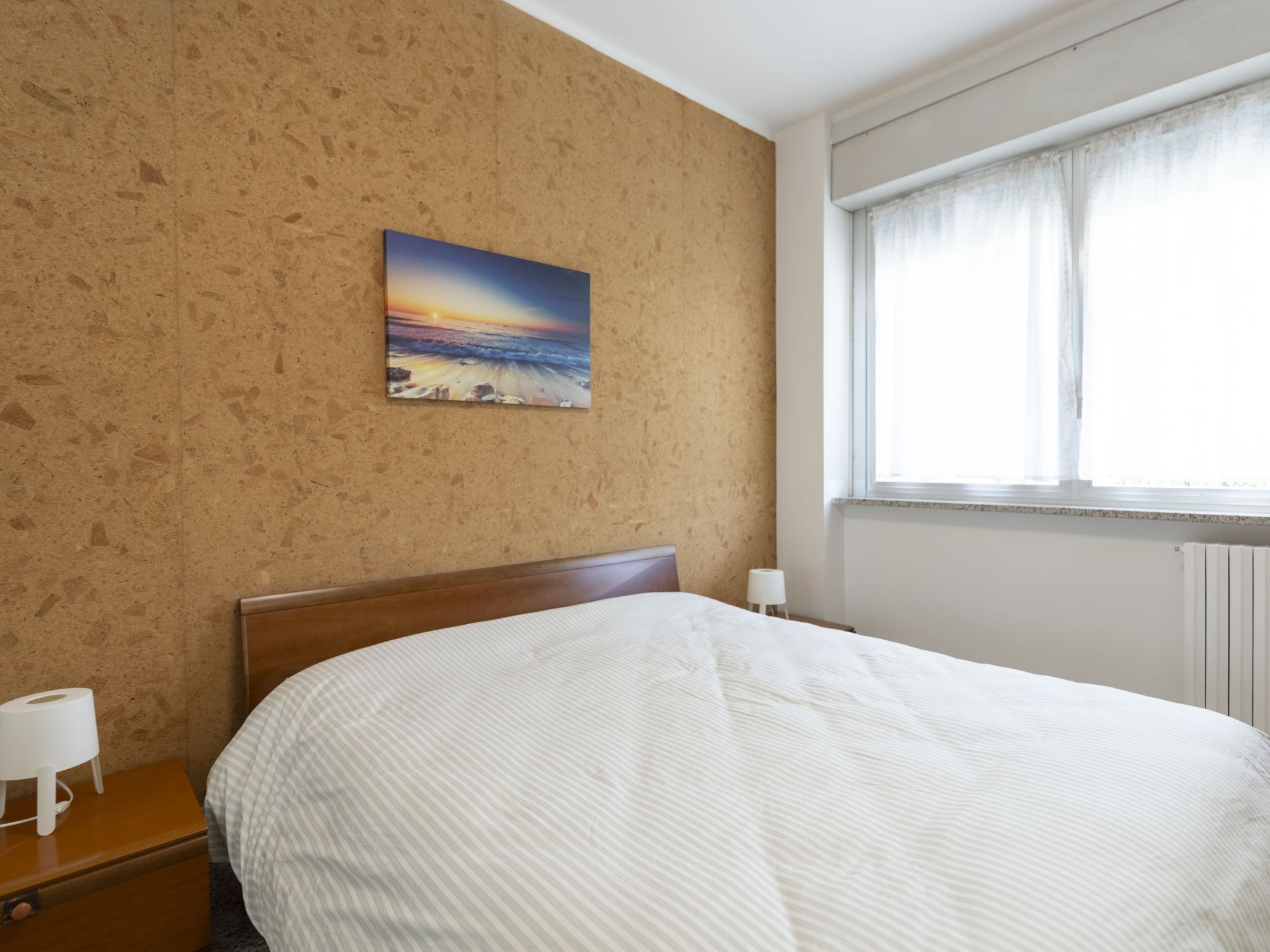 A hotel room with a bed, a nightstand, a painting of a sunset, and a window with curtains.
