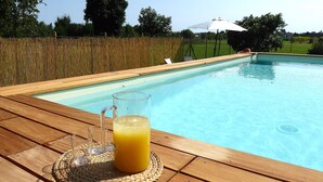 Eine Holzterrasse mit einem Glas Orangensaft und einem Glas Wasser, mit Blick auf einen Swimmingpool.