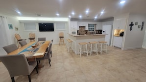 Una cocina moderna con armarios blancos, una isla central con taburetes y un comedor con una mesa larga y sillas.