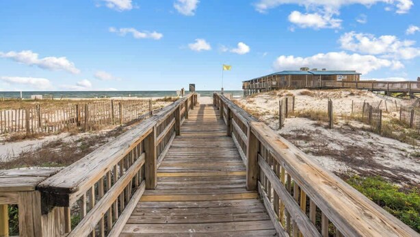 A wooden boardwalk leading to a beach area with a building in the background. - Boardwalk 584 (Gulf Shores)