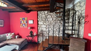 Un salon chaleureux avec un mur en pierre, un plafond en bois, un mur rouge, un canapé, une petite table avec une lampe et un fauteuil en osier.