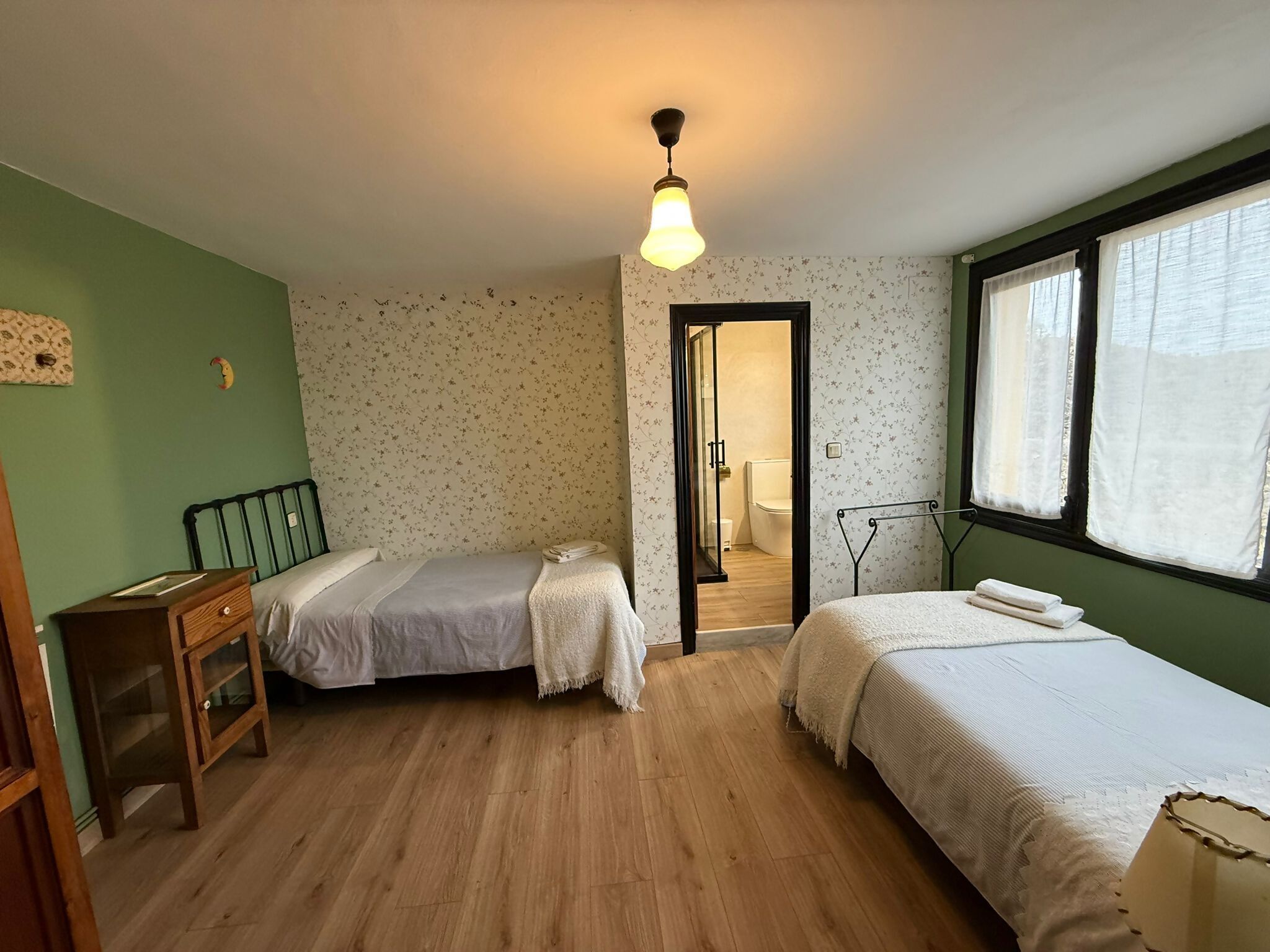 Una stanza con due letti, pavimento in legno, parete verde, una finestra con tende e una luce a soffitto.