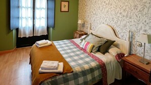 Un lit bien fait, avec une couvre-lit à carreaux, des oreillers et des serviettes pliées. On aperçoit une table de chevet avec une lampe et un tableau encadré accroché au mur.