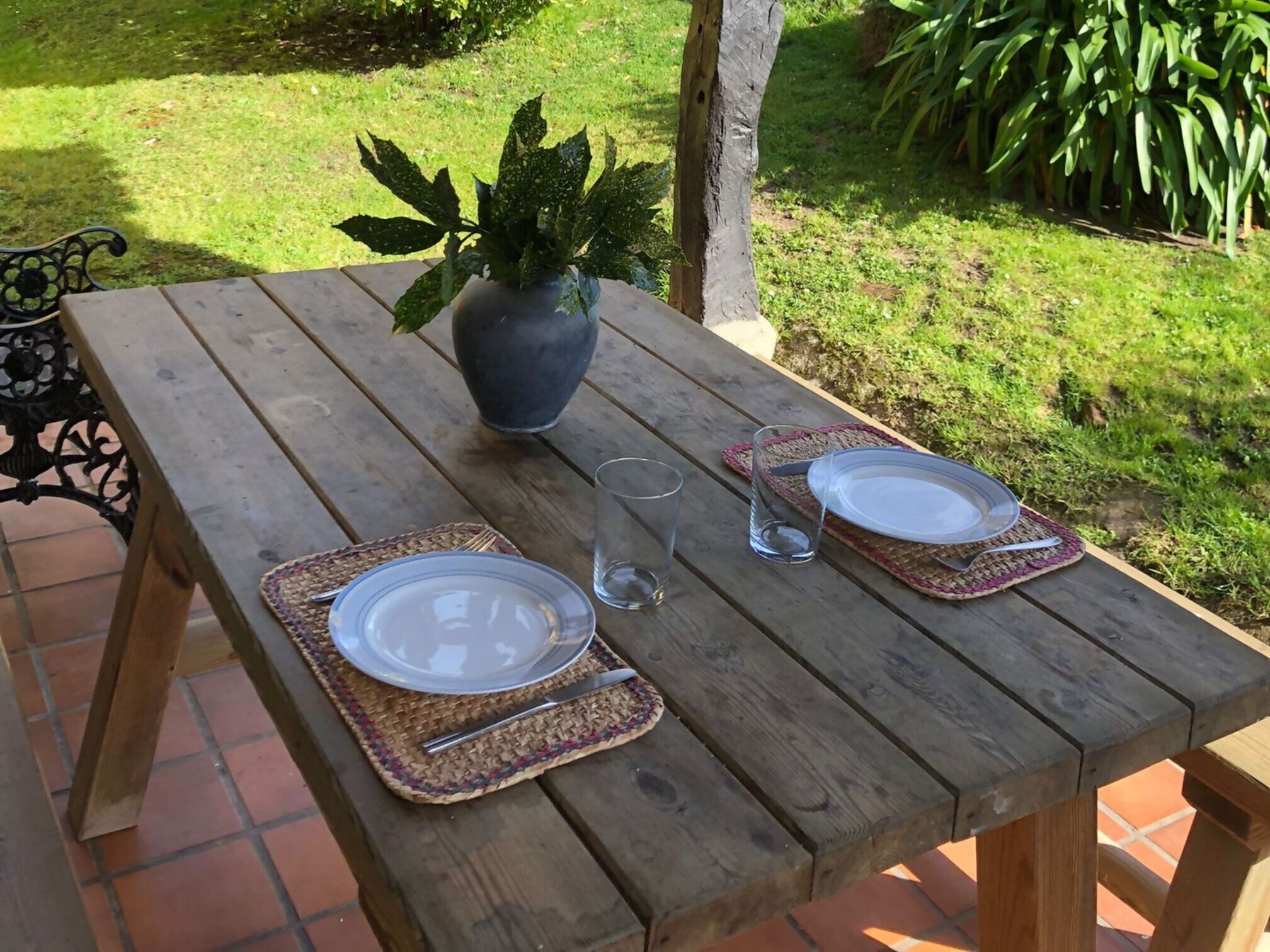 A wooden picnic table set with two plates, cutlery, and glasses, surrounded by greenery.