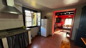 Une cuisine de taille réduite, équipée d’une cuisinière, d’un évier et d’une fenêtre. On aperçoit à l’arrière-plan un coin repas avec une table et des chaises en bois.