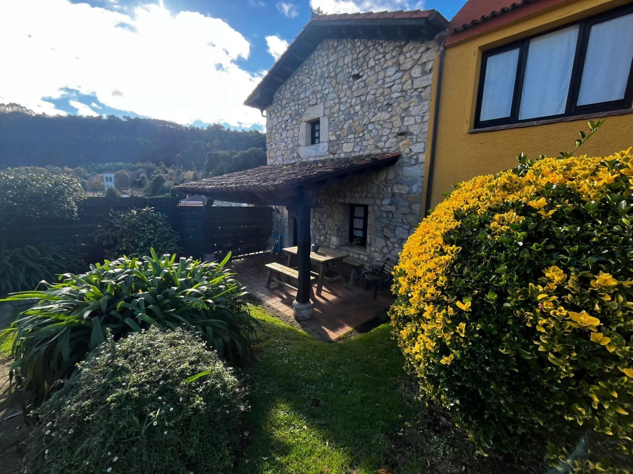 Una casa de pedra amb un pati, envoltada de vegetació abundant i un cel clar.