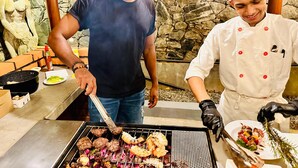 Zwei Personen grillen im Freien, im Hintergrund eine Steinmauer.