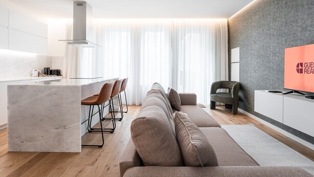 A modern living room with a sofa, a TV, and a kitchen area with a marble island and bar stools. - GuestReady - Little Johnny Stay (Porto)