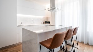 A modern kitchen with a central island, bar stools, and a large window with sheer curtains. - GuestReady - Little Johnny Stay (Porto)