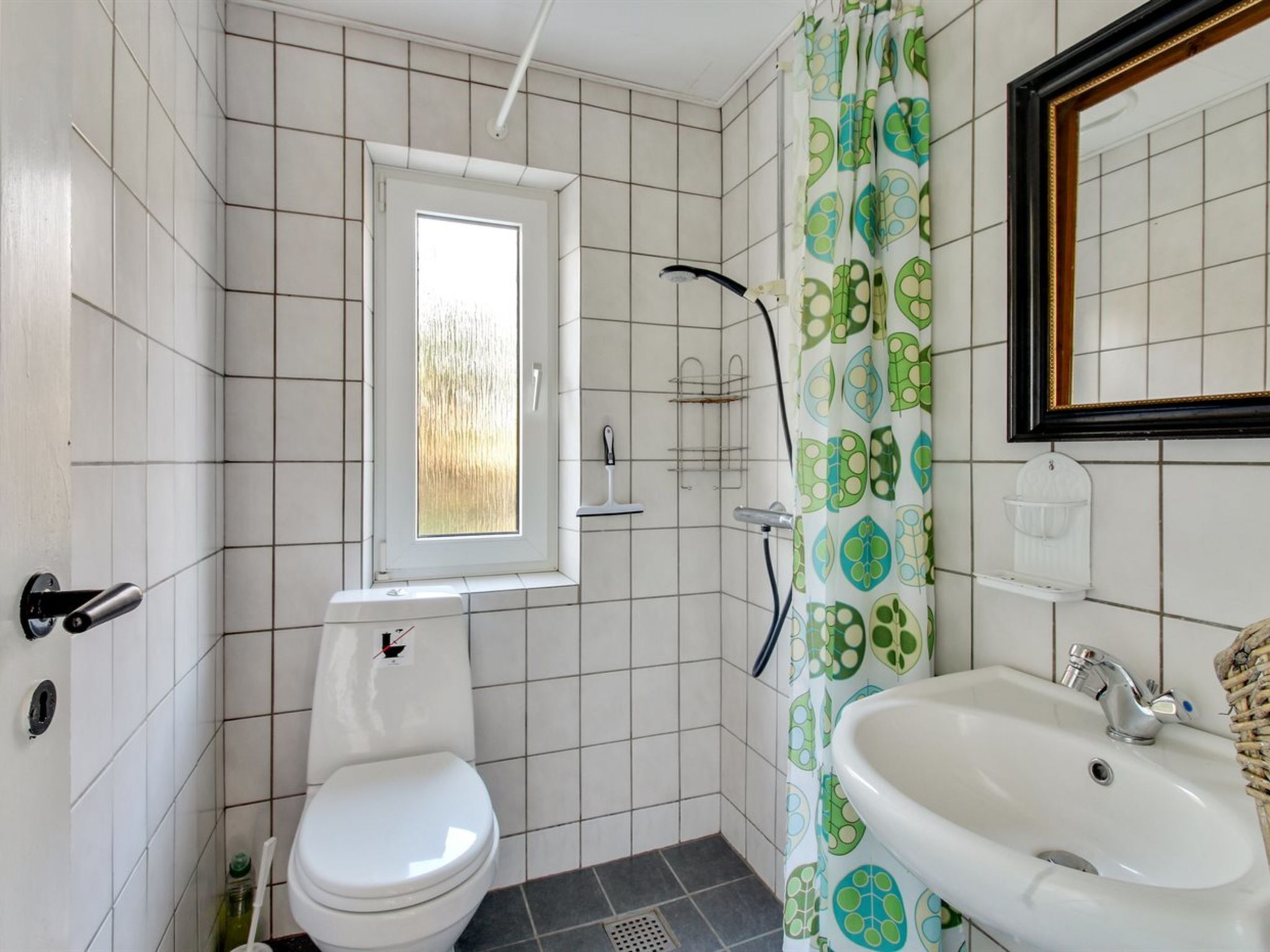 Une salle de bain avec des carreaux blancs, des toilettes, une douche, un lavabo et une fenêtre donnant sur un beau paysage.
