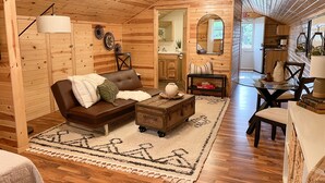 A cozy cabin interior with a wooden floor, a brown leather sofa, a small table with a vase, and a dining area with a chair and table. - The Dens at Bear Rock 2 with stairs access (Custer)