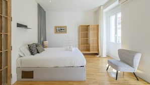 A modern hotel room with a large bed, a wooden wardrobe, a chair, and a window with curtains.