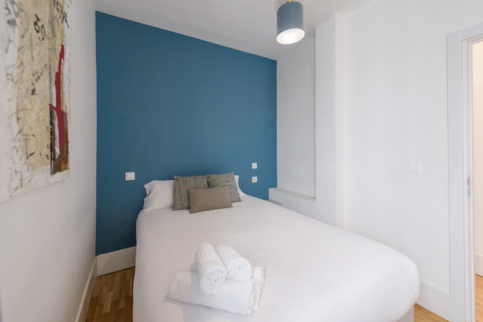 A bedroom with a bed, blue accent wall, white bedding, and a painting on the left.