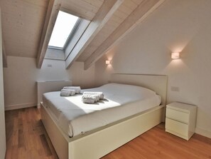 Ein Schlafzimmer mit schrägverlaufender Decke, einem Oberlicht, einem Bett mit weißer Bettwäsche, einem Nachttisch und einem Beistelltisch.