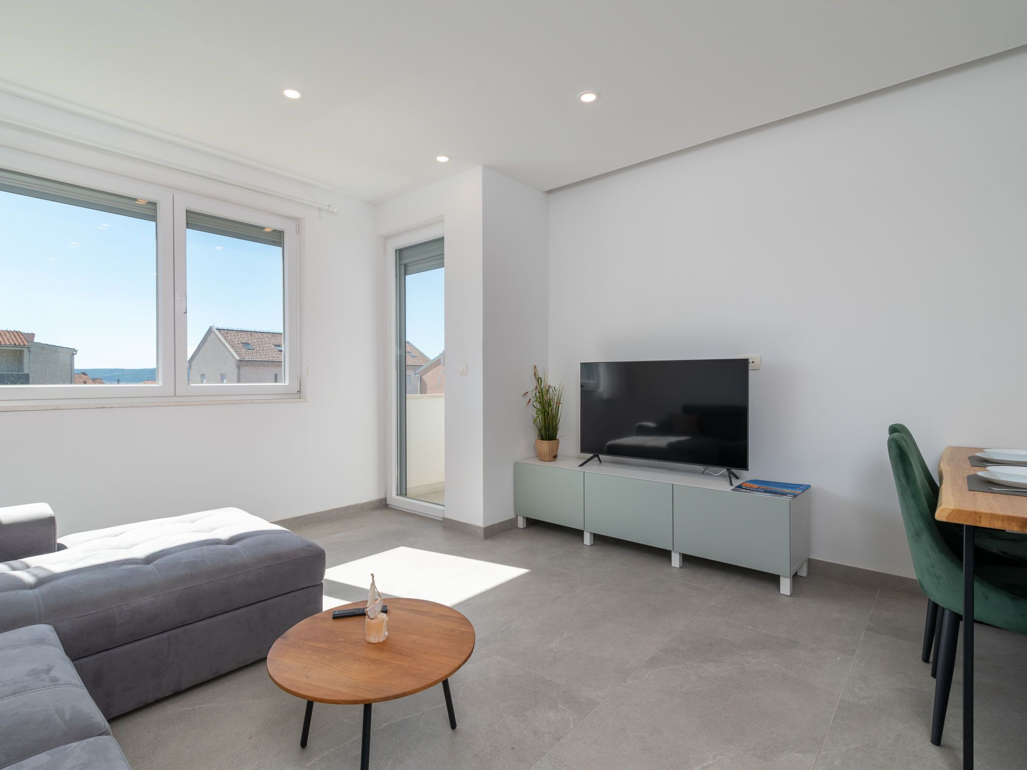 Una sala moderna con un sofá gris, una mesa de centro de madera, un televisor de pantalla plana sobre un mueble bajo y un rincón comedor con una silla verde.