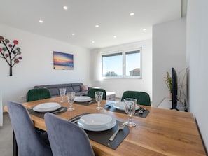 Un coin repas avec une table en bois, dressée pour quatre personnes, entourée de chaises. On y trouve également un canapé et une fenêtre donnant sur l’extérieur.