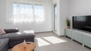 Un salon moderne avec un canapé gris, une table basse en bois, un téléviseur à écran plat posé sur un meuble vert clair, et de grandes fenêtres ornées de rideaux légers.