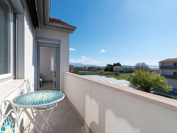 Un balcon avec une table et des chaises carrelées, une porte donnant sur une pièce intérieure, et une vue sur un quartier résidentiel avec des maisons et un ciel dégagé.