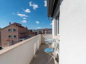Un balcon avec une balustrade blanche, une petite table agrémentée d’un coussin bleu et une vue sur les immeubles voisins.
