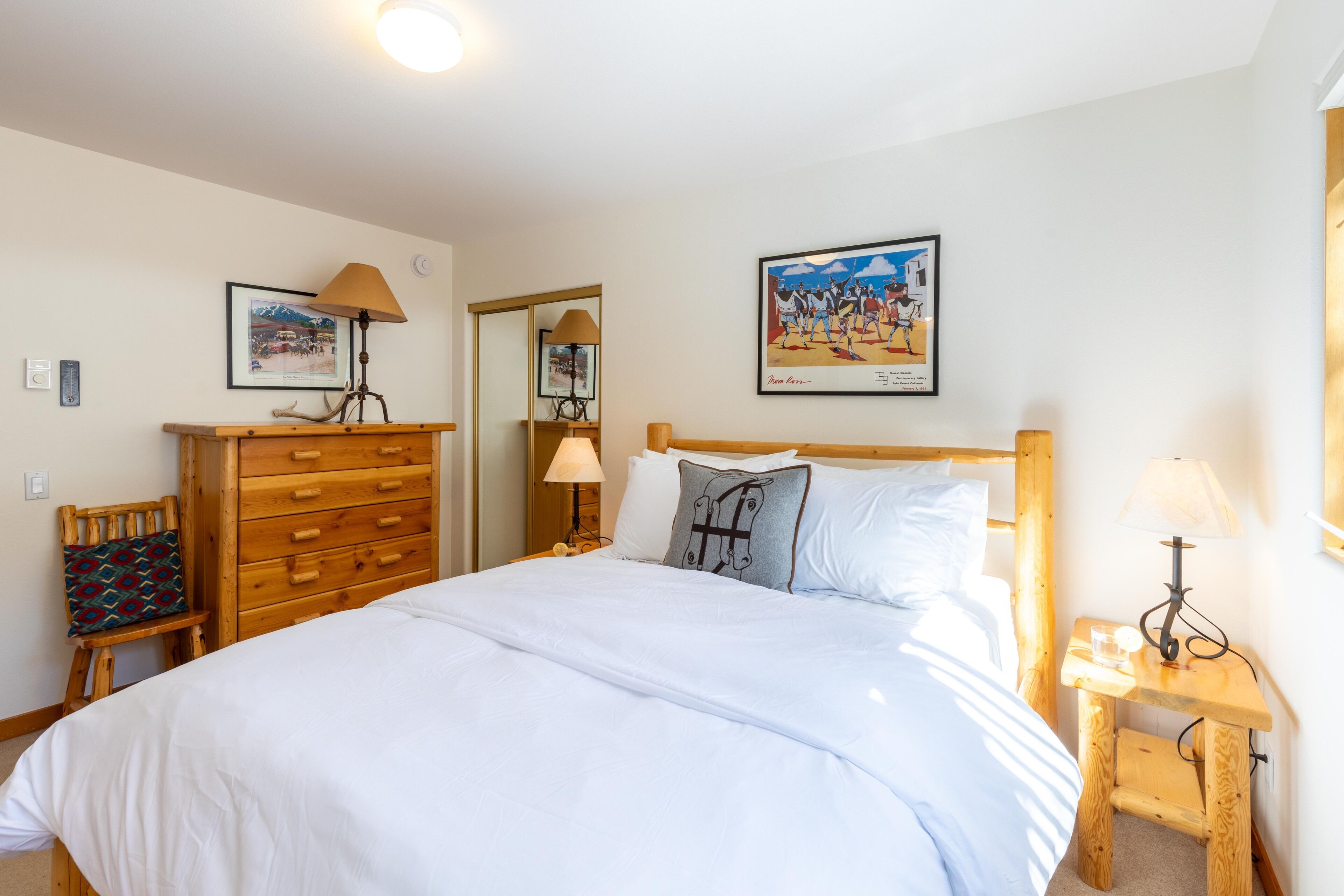 A bedroom with a large bed, wooden dresser, and nightstands with lamps. There are two framed pictures on the wall.