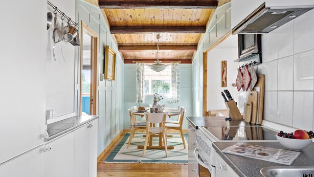 Une cuisine avec des poutres en bois, des meubles blancs, un coin repas avec une table et des chaises, et une fenêtre avec des rideaux.