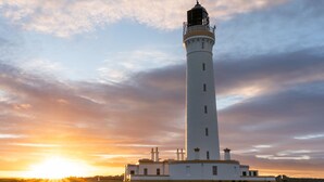 Un alto faro blanco con un balcón, silueteado contra un cielo al atardecer.