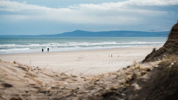 Una playa con una persona caminando, una orilla arenosa y una montaña a lo lejos.