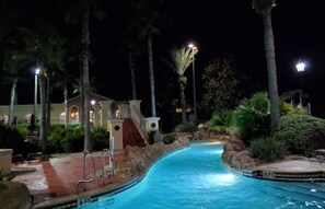 Un espace piscine nocturne avec un chemin rouge menant à la piscine, entouré de palmiers et d’un bâtiment dont l’entrée est illuminée.