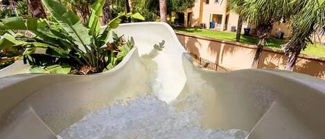 Un tobogán de agua con piscina en la parte inferior, rodeado de palmeras y un edificio al fondo.