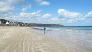 Une vaste plage de sable fin, avec des vagues douces, un ciel dégagé et des collines qui se dessinent à l’horizon.