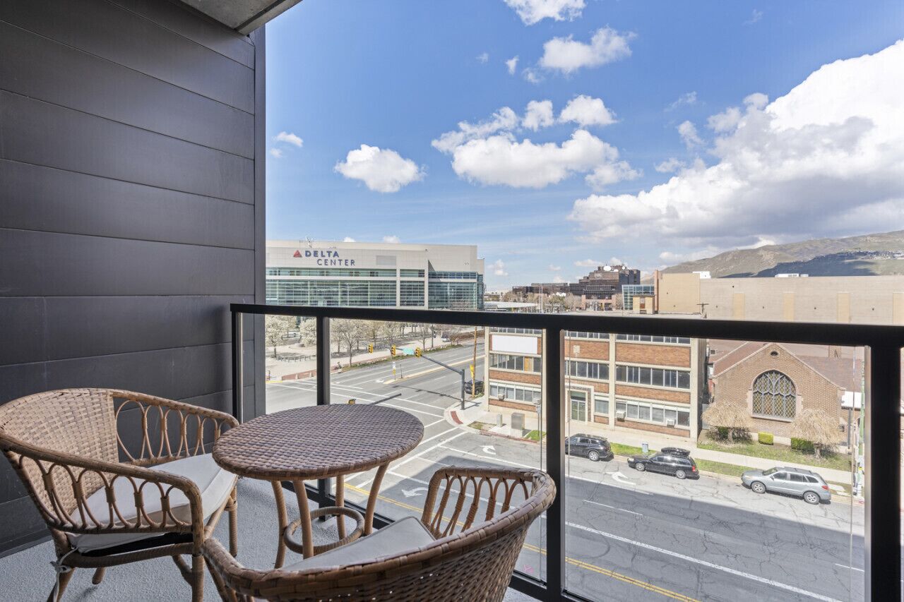 A balcony with wicker chairs and a table, overlooking a street with buildings, including a Delta Center.