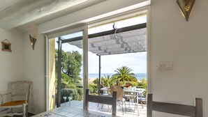 Une pièce avec un sol carrelé, une table entourée de chaises, une vue sur la plage et une porte donnant sur l’extérieur.
