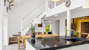 A modern kitchen with a black countertop, white cabinetry, and a stainless steel sink. There is a dining area with a wooden table and chairs, and a staircase leading to an upper level. - "Marrit" - 500m from the sea by Interhome (Glesborg)