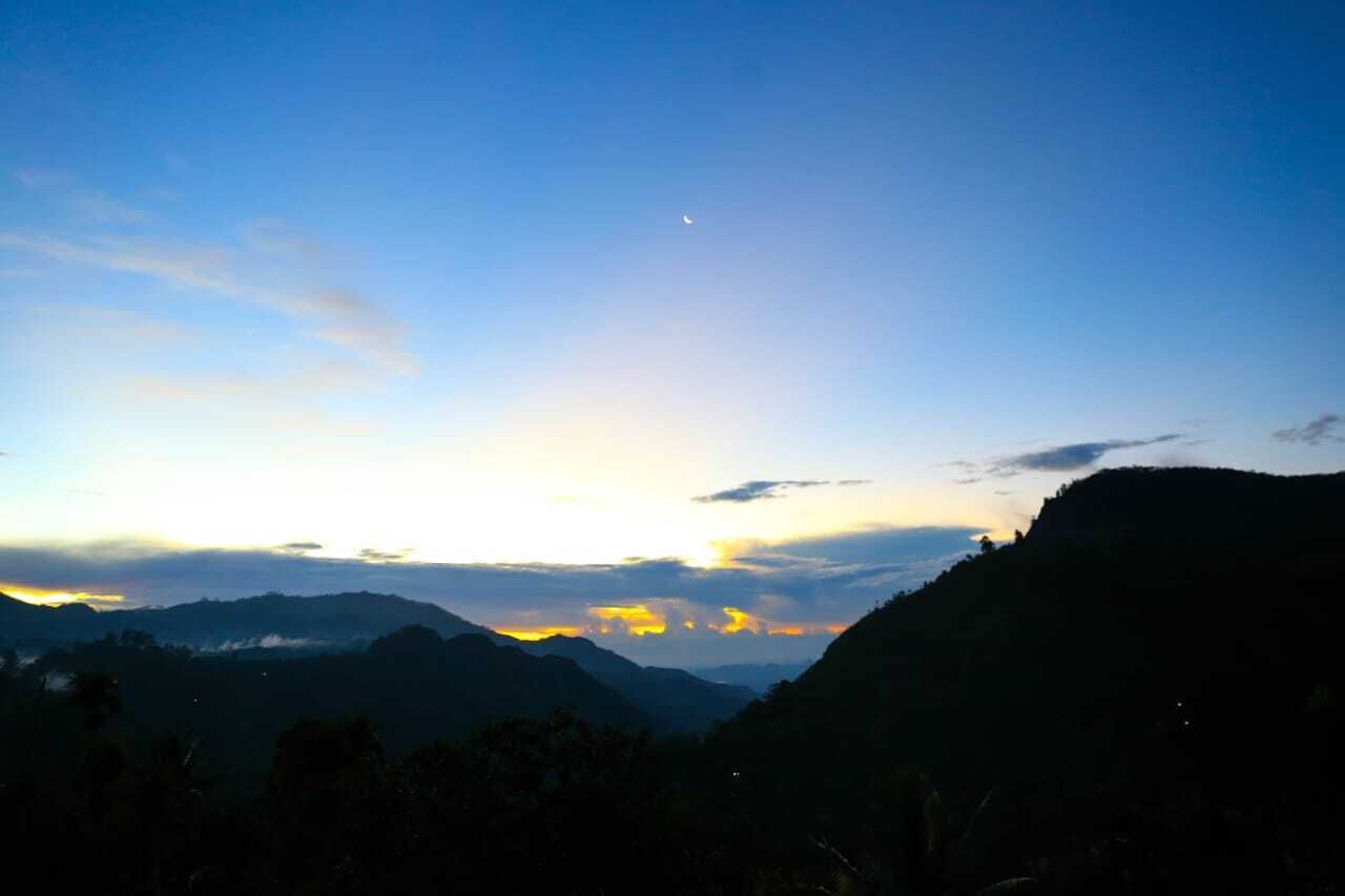 Paisagem montanhosa ao entardecer, com uma lua crescente visível no céu.