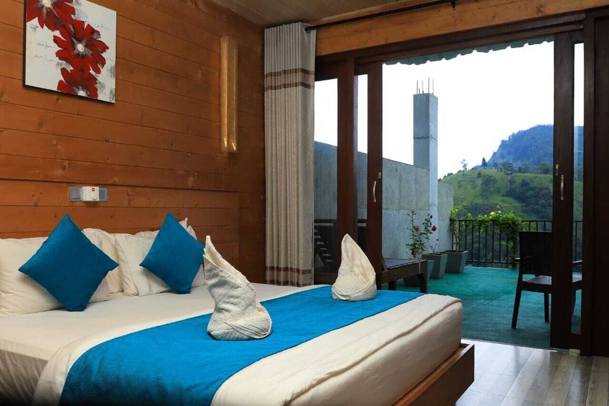 Ein Hotelzimmer mit Bett, blauen Kissen, einer blauen Bettdecke und Blick auf einen Balkon mit Pflanzen und einem Stuhl.