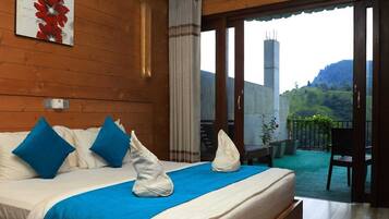 A hotel room with a bed, blue pillows, a blue bedspread, and a view of a balcony with plants and a chair.
