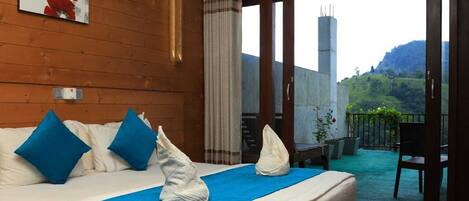 A hotel room with a bed, blue pillows, a blue bedspread, and a view of a balcony with plants and a chair.