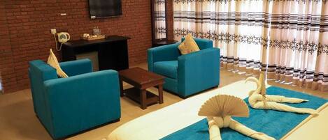 A living room with blue sofas, a wooden coffee table, a flat-screen TV mounted on a brick wall, a mirror, and a window with patterned curtains.