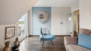Ein Wohnzimmer mit blauem Sessel, beigefarbener Couch, Wanddeko und einem gerahmten Kunstwerk.