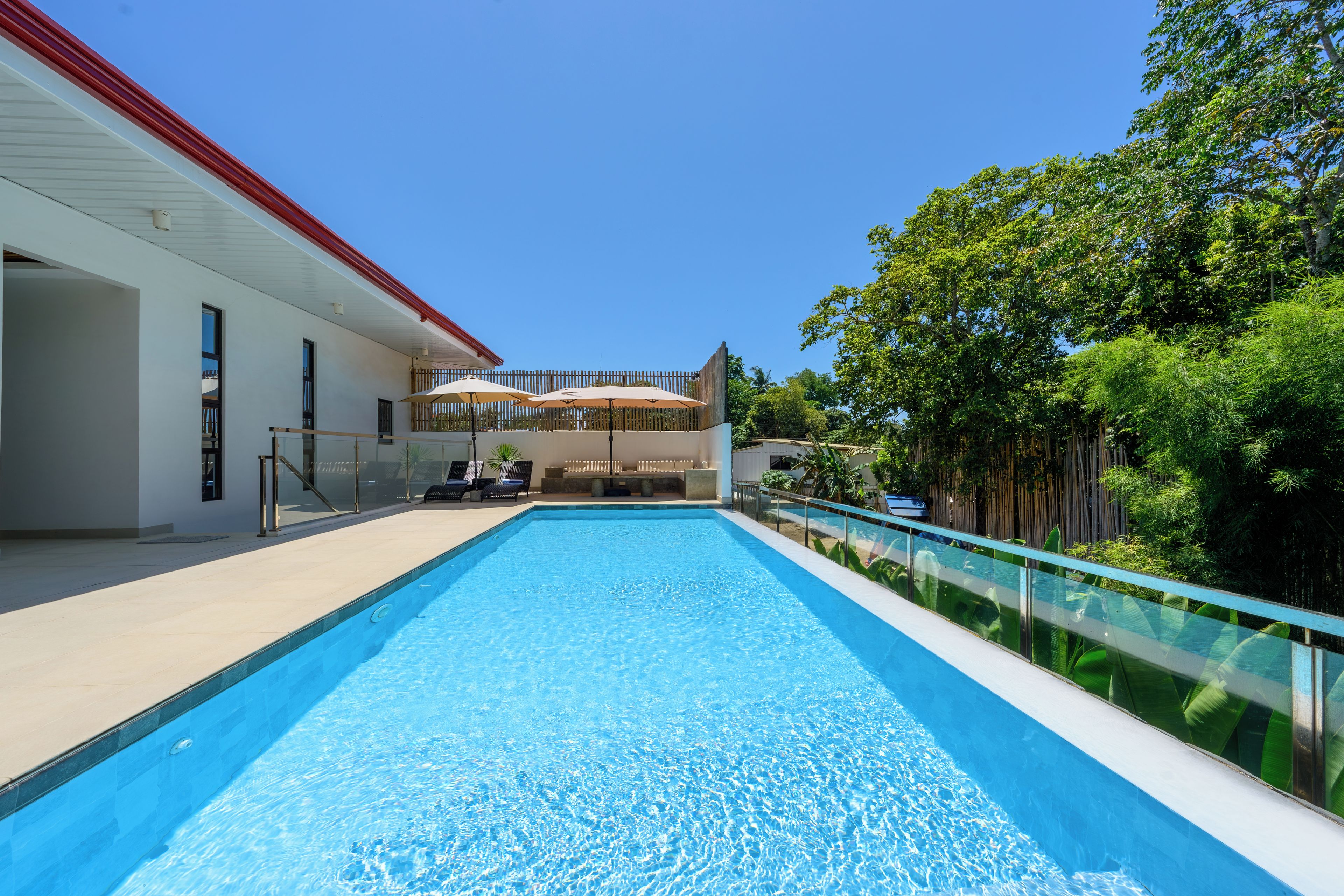 A swimming pool with a clear blue water surface, surrounded by a white building with a red roof, and lush green trees in the background.