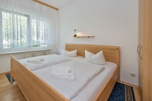 Uma cama bem arrumada com lençóis e toalhas brancas, uma mesa de cabeceira com abajur e uma janela com cortinas leves.