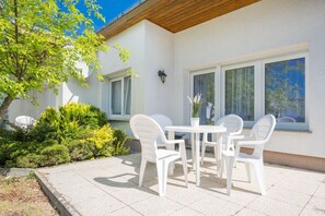 Eine Terrasse mit einem weißen Tisch und Stühlen, umgeben von Grünpflanzen und einem Gebäude mit großen Fenstern.